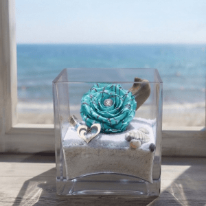 Rose éternelle de couleur bleu aqua présentée dans un élégant vase en verre avec un paysage marin en arrière-plan