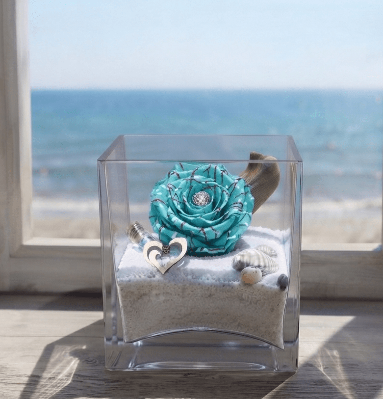 Rose éternelle de couleur bleu aqua présentée dans un élégant vase en verre avec un paysage marin en arrière-plan